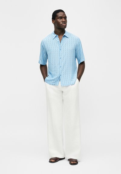 Άνδρας στέκεται με τα χέρια στις τσέπες φορώντας φωτεινά μπλε πουκαμισάκι με μοτίβα κοντομάνικο, λευκό παντελόνι με φαρδιά πόδια και μαύρα σανδάλια μπροστά σε λευκό φόντο.