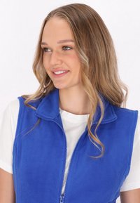 Blauer Fleece-Weste mit hohem Kragen und durchgehendem Reißverschluss. Sie verfügt über eine weiche Textur und ein nahtloses Design, getragen über einem weißen T-Shirt.