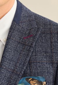 Marineblauer Tweed-Blazer mit Karomuster; verfügt über einen violetten Akzent am Revers und ein Einstecktuch mit Schmetterlingen auf türkisfarbenem Hintergrund.