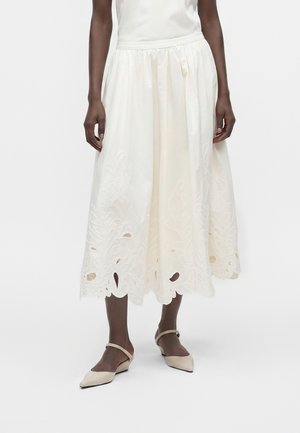 Mannequin féminin portant une jupe midi blanche avec broderie florale et ourlet festonné, associée à des chaussures beiges à bout pointu avec brides à la cheville.