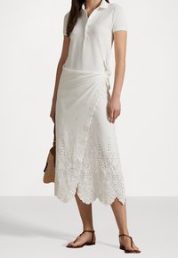 Biała bawełniana koszulka polo połączona z białą spódnicą w kształcie owijanej, zdobioną wzorami w oczka i falbaniastym dołem. Brązowe sandały i pleciona torba dopełniają całość stylizacji.