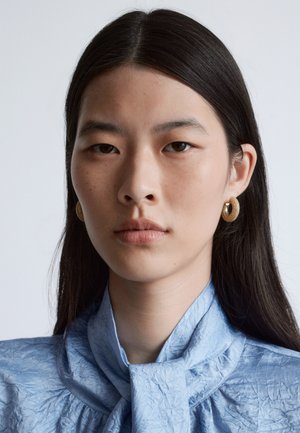 Goldene Creolen mit strukturierter Oberfläche, getragen zu einem hellblauen, zerknitterten Stoffhemd mit gebundenem Kragen. Glattes, gerades schwarzes Haar.