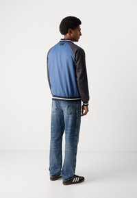 Blaue Bomberjacke mit schwarzen Ärmeln, gerippten Bündchen und Kragen; getragen mit hellblauen, geraden Jeans und braunen Sneakers.