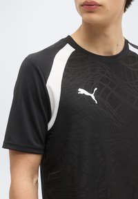 Νεαρό άτομο που φοράει μαύρο αθλητικό μπλουζάκι Puma με λευκές λεπτομέρειες στους ώμους και διακριτικό υφασμάτινο μοτίβο στο μπροστινό μέρος.