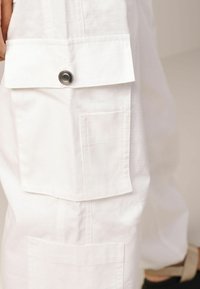 Vêtement blanc en tissu texturé, avec plusieurs poches et un détail à pression sur la partie supérieure des poches.