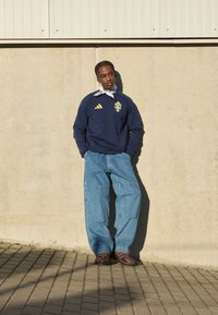 Temno moderna majica s kapuco z rumenim logotipom Adidas in grbom Švedske, v kombinaciji s svetlo modrimi ohlapnimi kavbojkami in rjavimi čevlji. Siva bandana okoli vratu.