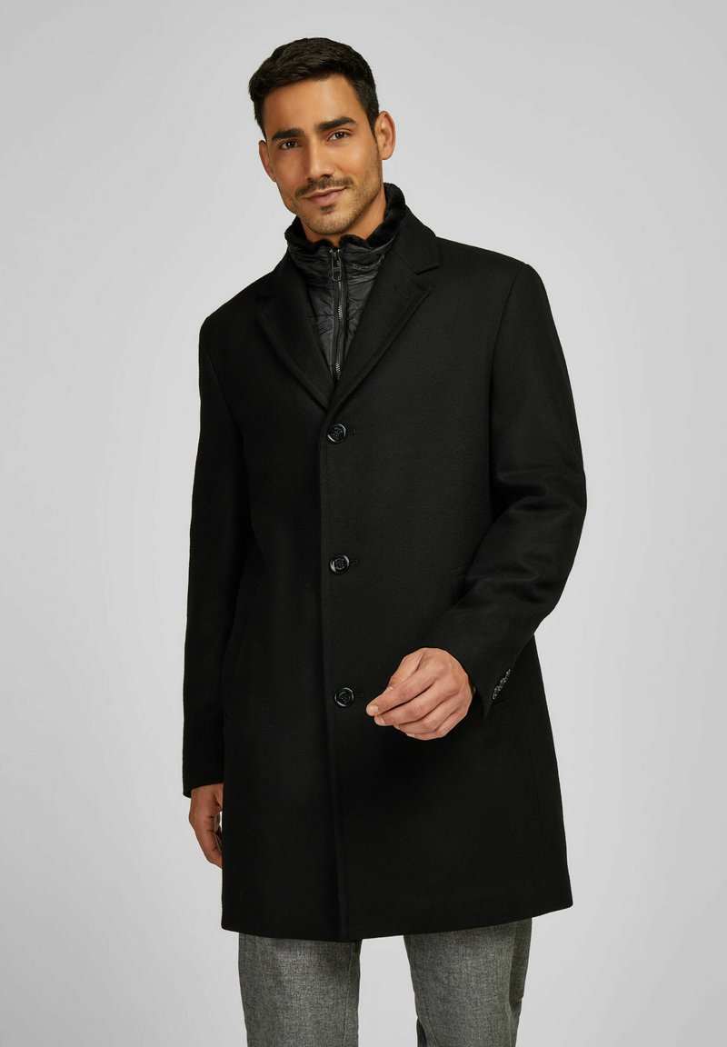 Cappotto di lana nero con colletto a scialle, sei bottoni e una consistenza liscia, indossato sopra uno strato interno trapuntato, abbinato a pantaloni grigi.