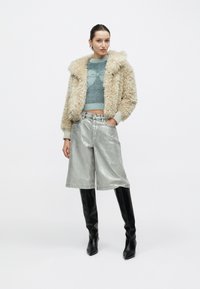 Kunstpelzjacke in Beige, hellblauer Strickpullover, metallisch silberne Shorts und schwarze kniehohe Stiefel; zeitgemäßes, strukturiertes Design.