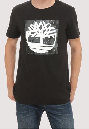 Homme portant un t-shirt noir avec un graphique d’arbre géométrique blanc à l’intérieur d’un carré sombre sur la poitrine, assorti à un jean bleu.