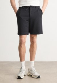 Shorts negri texturat, cu talie ajustabilă prin cordon, buzunare laterale și o croială obișnuită, asortați cu adidași groși gri și șosete albe.