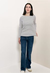 Lange mouwen gestreepte top in wit en marineblauw, gecombineerd met uitlopende blauwe jeans en witte sneakers, tegen een neutrale achtergrond.