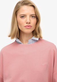 Różowy bawełniany sweatshirt z ribbanym dekoltem, z delikatnie haftowanym logo. Model nosi niebiesko-paskowaną koszulę pod spodem. Miękka faktura.
