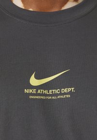 Šedé sportovní tričko s kulatým výstřihem. Na přední straně se nachází žluté logo Nike a text "NIKE ATHLETIC DEPT. ENGINEERED FOR ALL ATHLETES" pod ním.