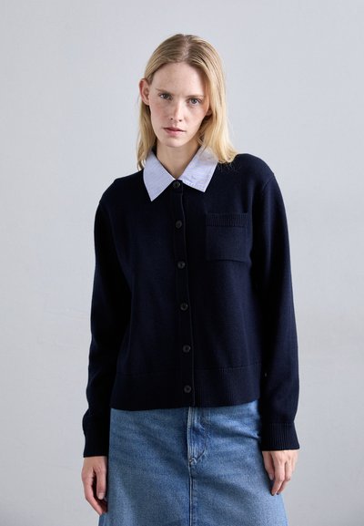 Cardigan navy con tasca, scollatura rotonda e chiusura con bottoni, indossato sopra una camicia colletto a righe azzurre. Texture in maglia morbida.