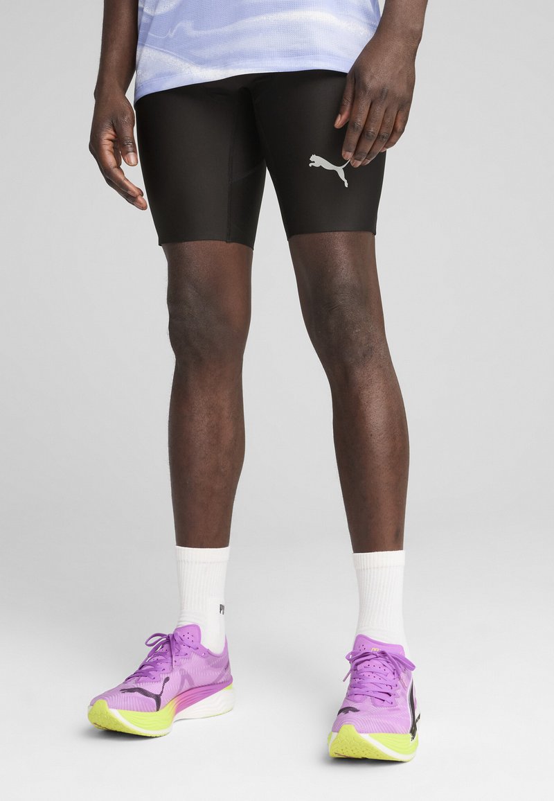 Sorte atletiske shorts med et PUMA-logo, parret med lilla og grønne løbesko. Hvite sokker fuldender det sporty look.