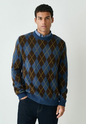 Sweater im Argyle-Muster in Blau, Braun und Gelb. Aus weichem Wollstrick, mit Rundhalsausschnitt, gerippten Bündchen und Saum. Über einem blauen Hemd mit Kragen getragen.