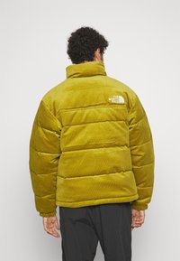 Pufferjacka i olivgrön, tillverkad av manchester. Har en hög krage och ribbstickade muddar. Visar The North Face-logotypen på övre delen av ryggen.