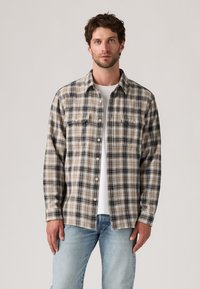 Rutet flanellskjorte i beige, svart og brunt. Har knappestolpe foran, to brystlommer og lange ermer. Brukt over en hvit T-skjorte.