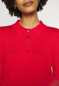 Rotes Strickpolo mit Kragen und fünf schwarzen Knopfverschlüssen. Hat lange Ärmel und eine weiche Textur, geeignet für legere Kleidung.