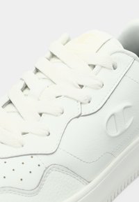 Λευκό δερμάτινο αθλητικό παπούτσι με υφασμάτινες πλευρικές πάνελ, στρογγυλές οπές στο μπροστινό μέρος, επίπεδα λευκά κορδόνια και ανάγλυφο λογότυπο στην εξωτερική πλευρά.