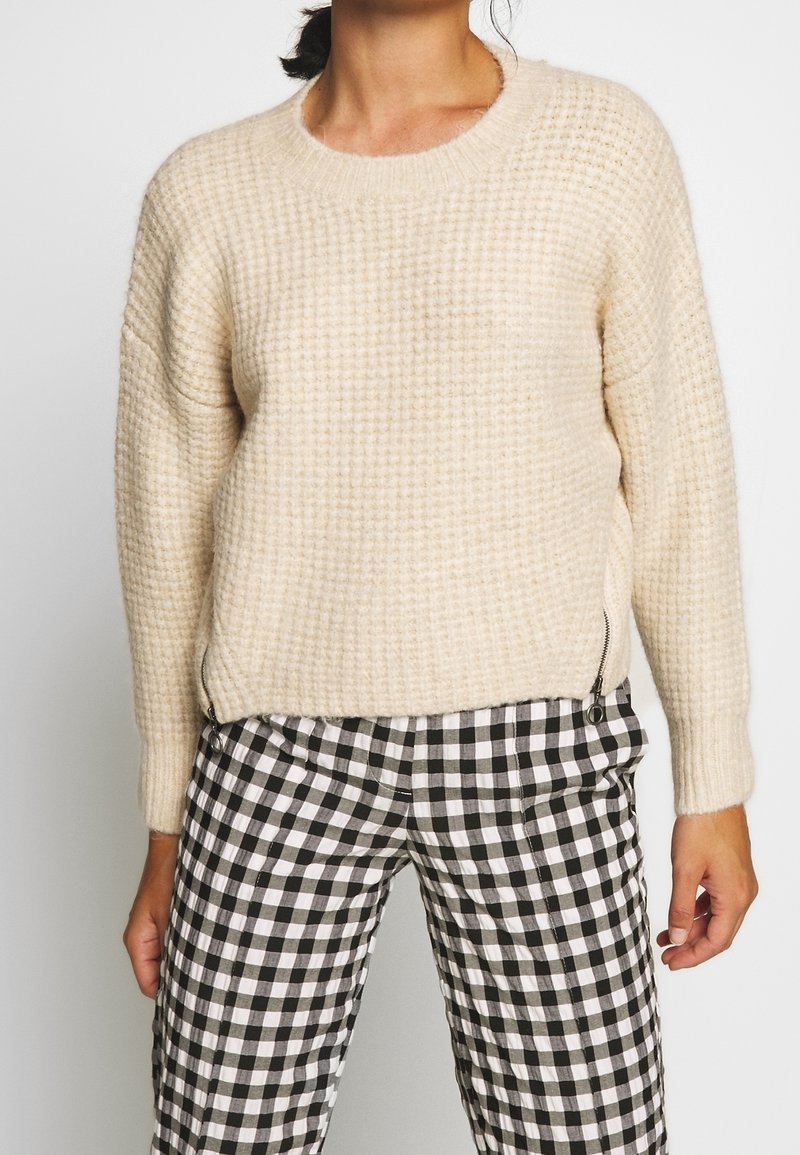 Pull en maille couleur crème avec un motif texturé, col rond et fermetures éclair latérales, assorti à un pantalon à carreaux noir et blanc.