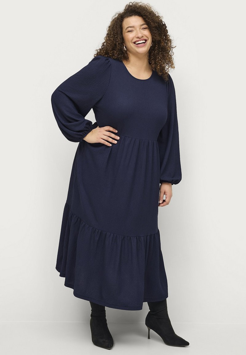 Vestido de manga larga color azul marino con un tejido texturizado, escote redondo y dobladillo con volantes en capas. Combinado con botas negras hasta la rodilla.