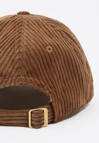 Gorra de pana marrón con textura de nervios verticales, que cuenta con una correa ajustable en tono dorado y un acento de logo metálico en la parte trasera.