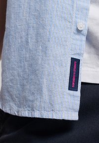 Camisa de manga corta a rayas azul claro en un tejido texturizado, con un botón blanco y una etiqueta azul con texto rosa en el dobladillo.