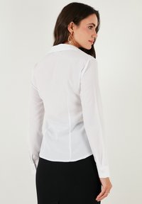 White long-sleeve blouse with a pointed collar, button cuffs, structured fit, and lightweight, smooth fabric. Back features subtle seams.