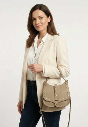 Vrouw in een beige blazer en wit overhemd met een beige leren handtas met studs over haar arm, tegen een effen achtergrond.
