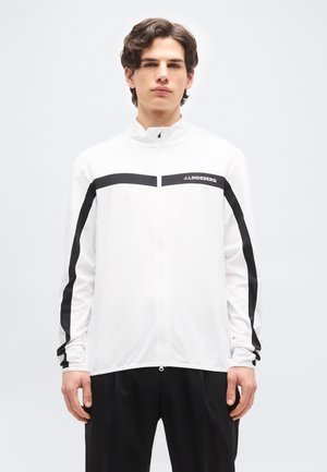 Jaunas vyras su bangeliais rudos spalvos plaukais dėvi baltą užsegamą striukę su juodomis juostelėmis ir užrašu „J.LINDEBERG“, derintą su juodomis kelnėmis.