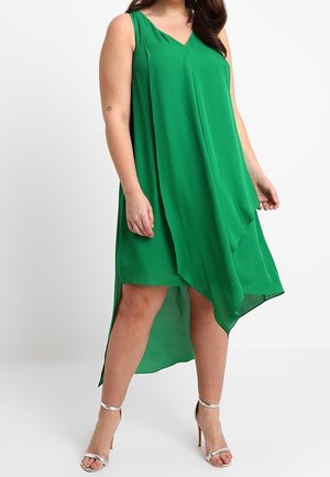 Woman wearing sleeveless green asymmetrical dress with high-low hem and silver high-heeled sandals, standing against a white background.