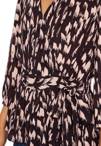 Schwarze und rosa gemusterte Bluse mit Wickeldesign, ausgestattet mit einem Peplum-Bund und einem gebundenen Taillendetail. Weicher Stoff mit fließender Textur.