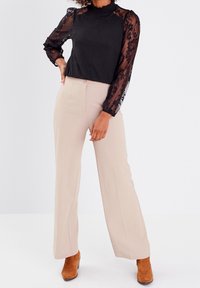 Top noir avec manches longues en dentelle et col montant ; associé à un pantalon beige cintré et des bottines marron. Tissu lisse, design élégant.