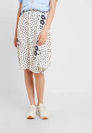 Kobieta w białej spódnicy do kolan w czarne kwiatowe i grochowe wzory, jasno-niebieskiej bluzce oraz białych tenisówkach.