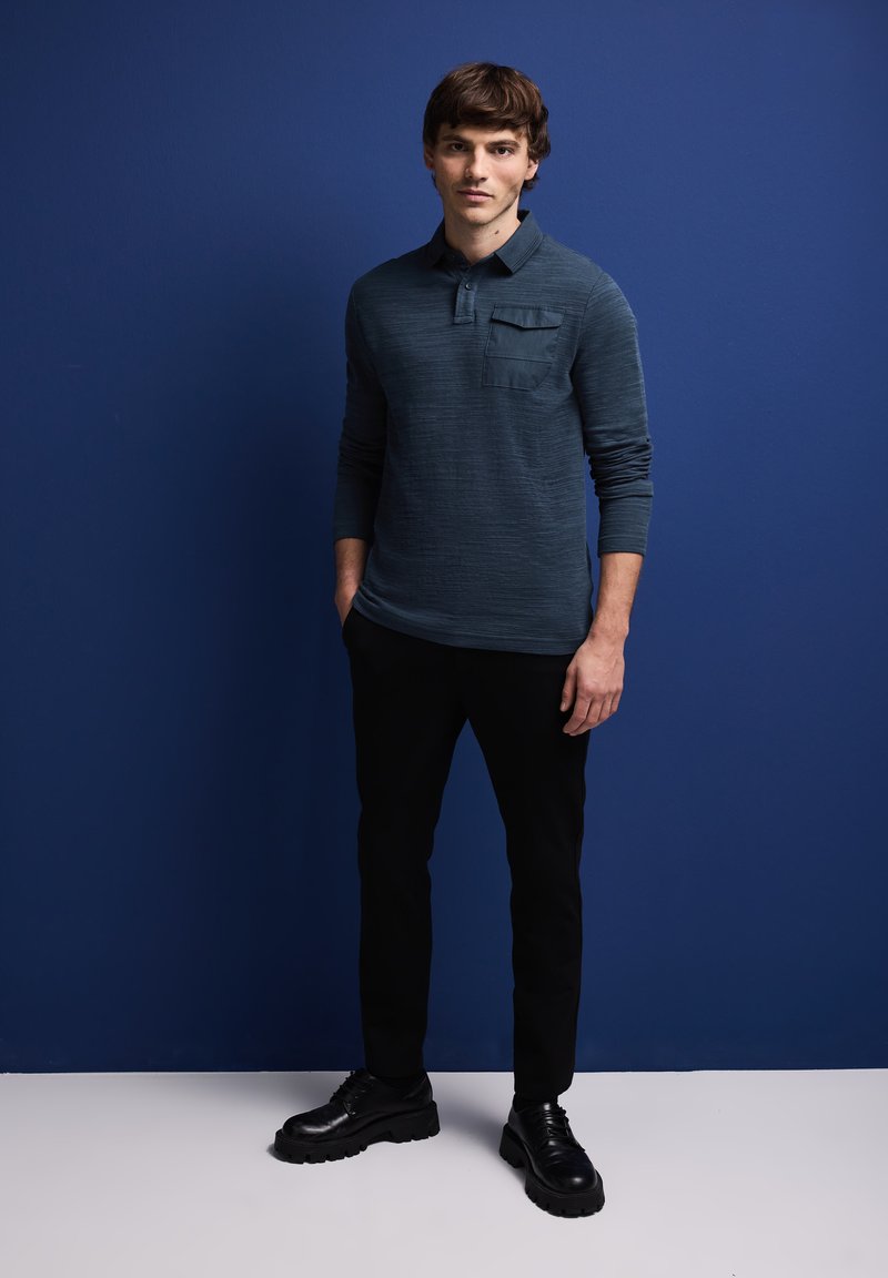 Tmavě modré dlouhé polo tričko s texturovanou tkaninou a přední kapsou, spárované s černými slim-fit kalhotami a černými šněrovacími botami.