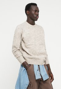 Beige textured sweater with a crew neck and ribbed cuffs, worn over brown cargo pants, with a light blue denim shirt tied at the waist.