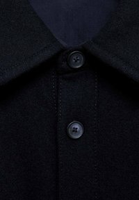 Gros plan sur le col d’un manteau en laine noire avec deux boutons noirs à quatre trous fixés sur la patte de boutonnage avant.