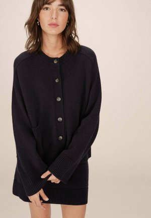 Cardigan bleu marine foncé avec un col rond, de grands boutons, des poignets côtelés et deux poches latérales, fabriqué à partir d'un tissu tricoté texturé.