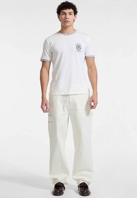 Biały t-shirt z krótkim rękawem z szarym obszyciem i logo, zestawiony z luźnymi kremowymi spodniami z kieszeniami i metalowymi elementami.