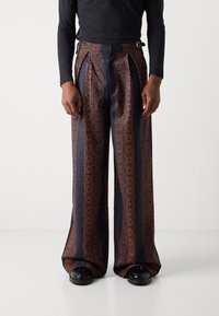 Pantalones de patrón navy con diseños geométricos naranjas, de pierna ancha, con pliegues y hebillas laterales. La tela parece suave y ligera.