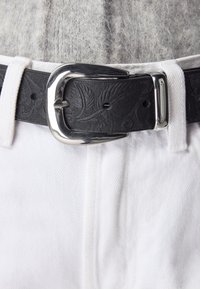 Czarny skórzany pasek z wytłoczonym wzorem liści i srebrną klamrą, noszony na białych spodniach i szarym dzianinowym swetrze.