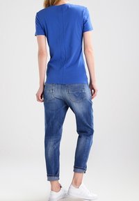 Kvinna i en blå kortärmad skjorta, uppvikta blå jeans och vita sneakers stående vänd bort från kameran mot en neutral bakgrund.