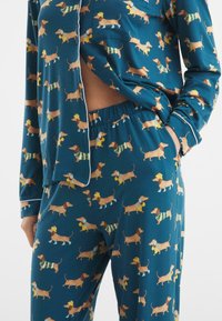Passendes Pyjama-Set aus teal-farbenem Stoff mit wiederholtem Dackel-Muster. Langärmeliges Oberteil mit Tasche; Hose mit elastischem Bund. Weicher, leichter Stoff.