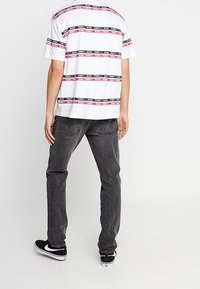 Camisa blanca de mangas cortas con rayas horizontales rojas y negras, con texto de logo. Combinada con jeans gris oscuro y zapatillas negras.