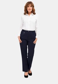 Witte overhemden met knopen, donkere geruite broek met zijzakken, metalen hardware accenten en zwarte schoenen. Model poseert tegen een neutrale achtergrond.