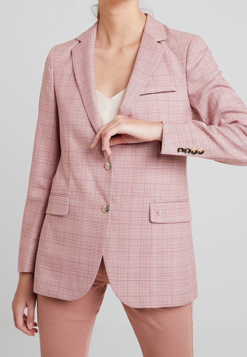 Femme portant un blazer rose à carreaux avec des boutons dorés sur un haut blanc et un pantalon rose, ajustant le bouton avant du blazer.