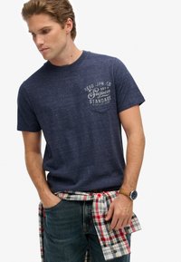 Hombre viste una camiseta gráfica azul marino, jeans azules y una camisa de cuadros atada a la cintura, mirando hacia abajo con las manos cerca de los bolsillos.