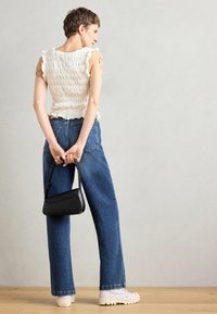 White smocked top with ruffled edges, high-waisted blue jeans, black handbag, and white sneakers. Model stands with back turned.