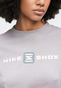 Šedé bavlnené tričko s okrúhlym výstrihom a stredovým potlačeným logom, na ktorom je bielymi a modrými písmenami napísané "NIKE SHOX".